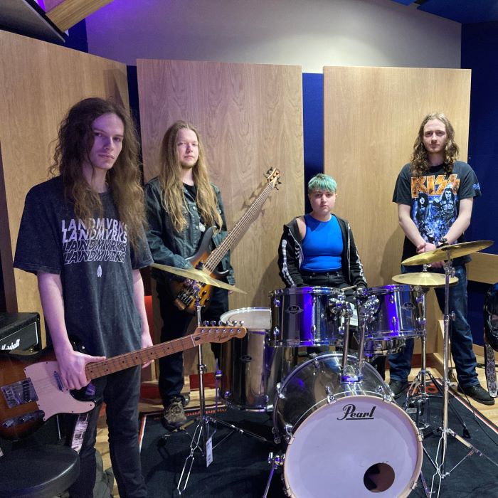 Five piece rock band in studio. Four long-haired young man and girl drummer with short hair
