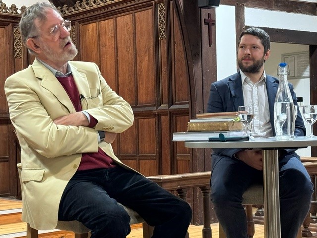 older man in cream jacket and younger bearded man in grey suit on stools