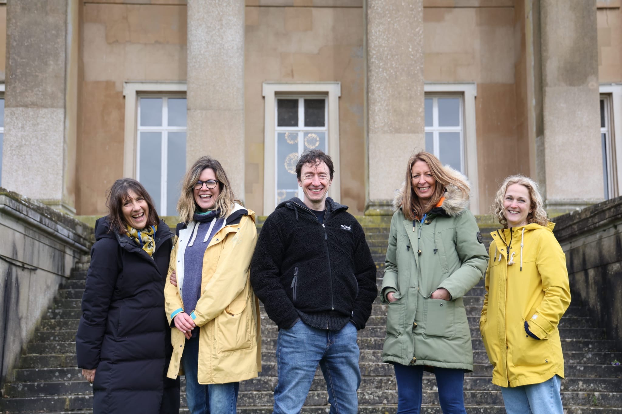Line up of five people,  four women and one man, outside large stately home