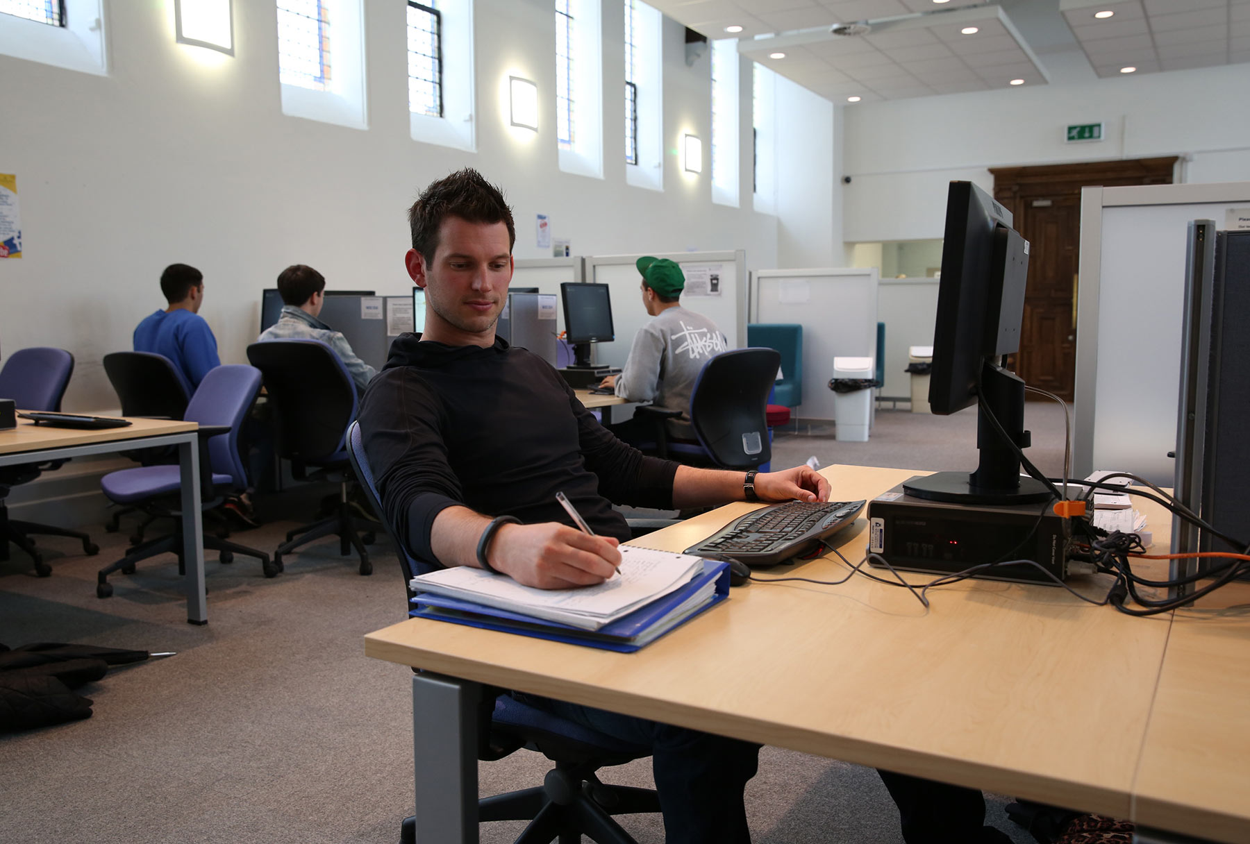 Student sitting at a computer station making notes on a notepad