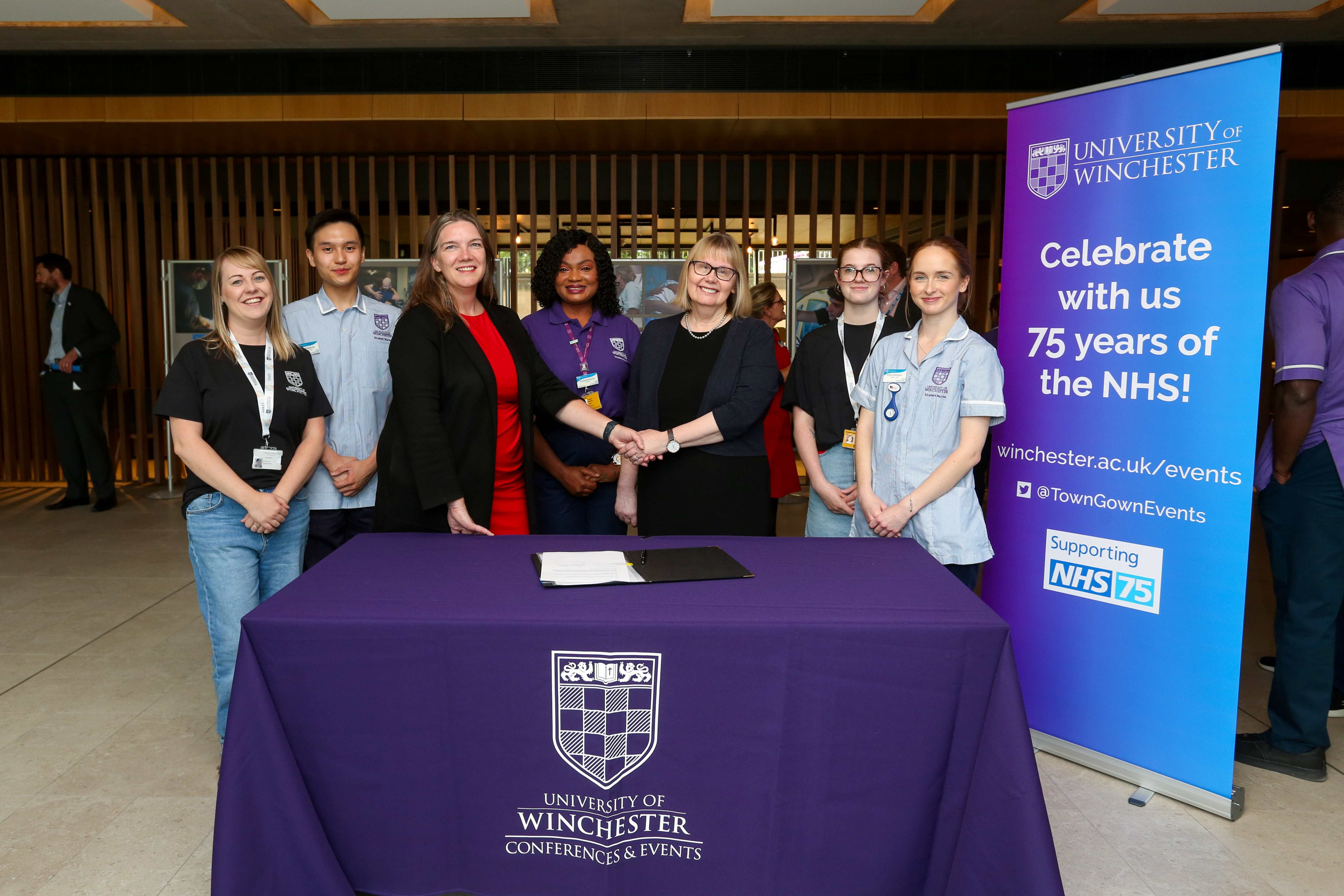 Alex Whifield, HHFT Chief Executive, shaking hands with Professor Sarah Greer, Vice Chancellor, in front of the partnership agreement, accompanied by five HWB students.
