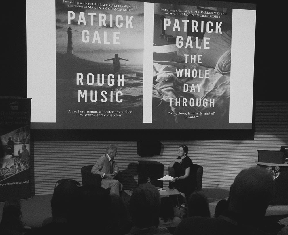 Two people sitting on chairs on stage talking with backdrop of book covers on an overhead screen