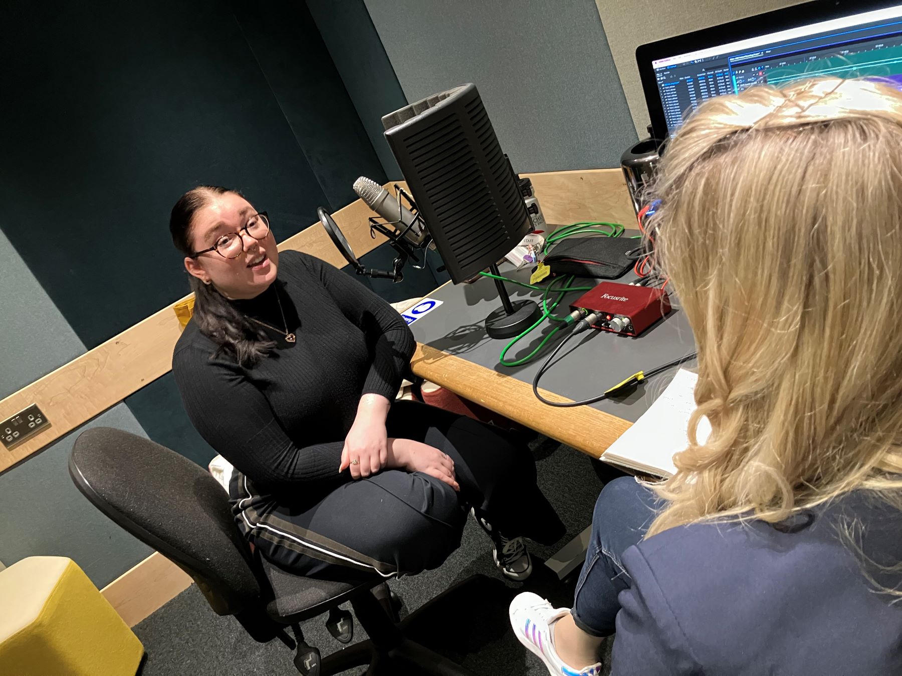 Two women in recording studio. One with dark hair and glasses faces camera. Blond woman with back to camera