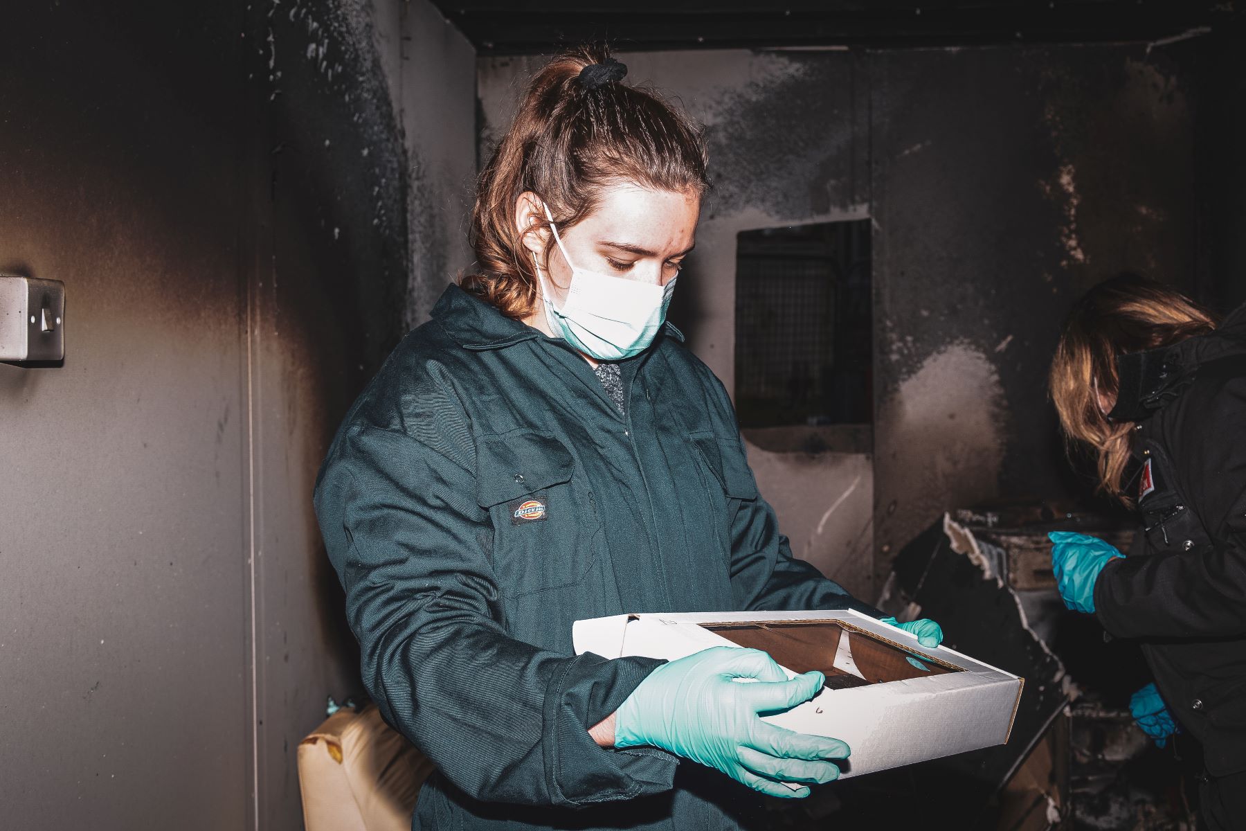 Girl on overalls and face mask carrying a tray at blackened fire scene