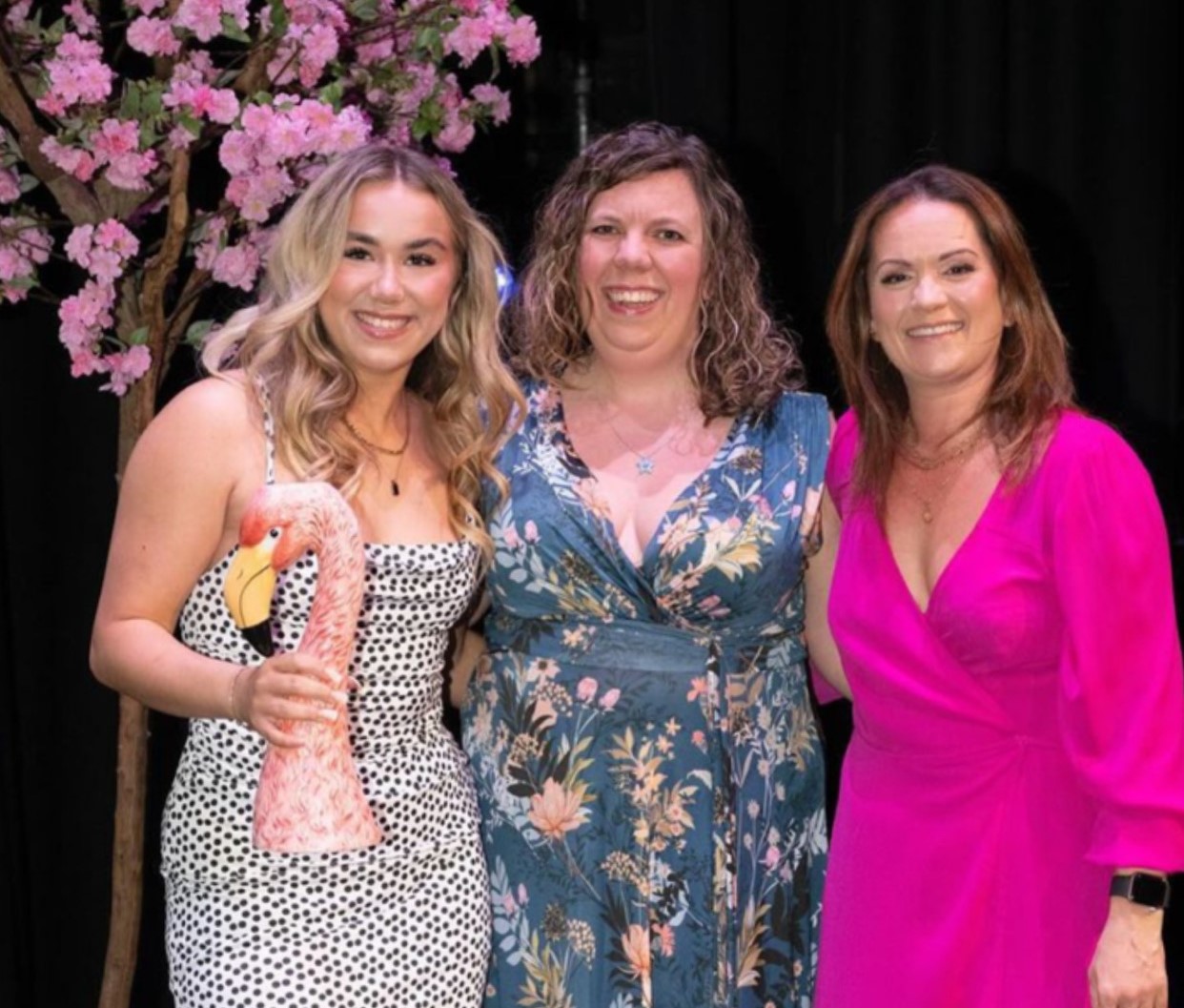 Three women in posh frocks. Youngest on left has trophy shaped like pink flamingo