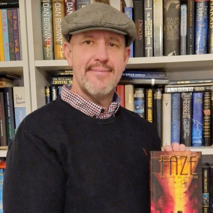 Man with cap holding book in front of bookcase