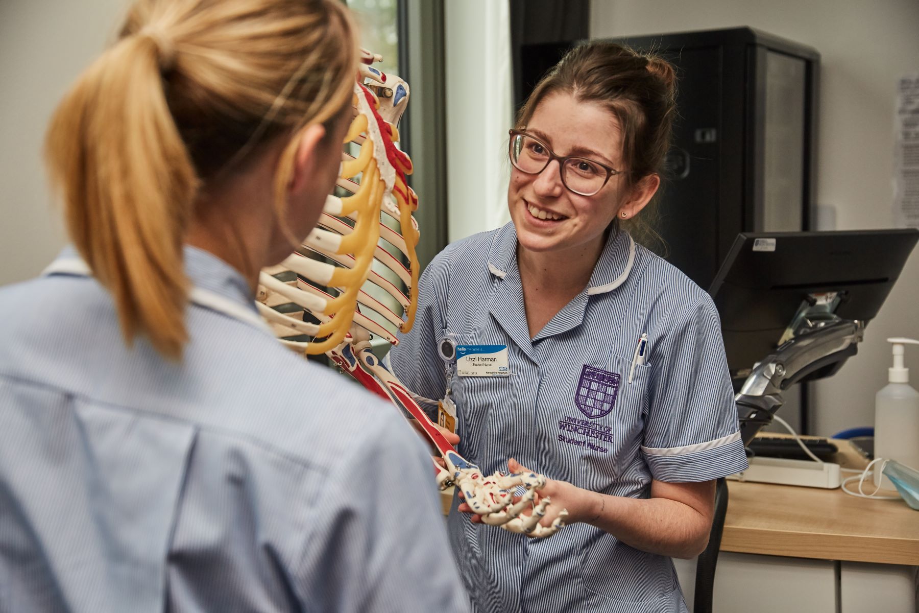 Two female student nurses talking next to skeleton. One has back to camera