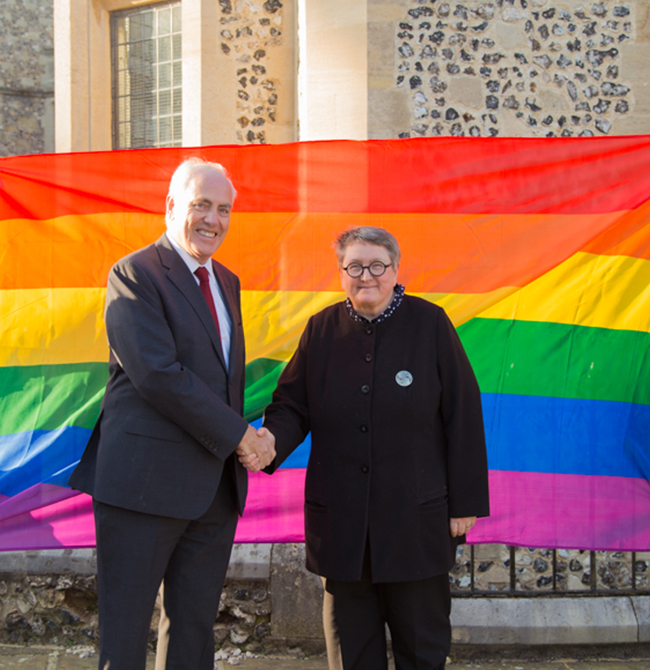 Two people shaking hands in front of a rainbow Pride flag