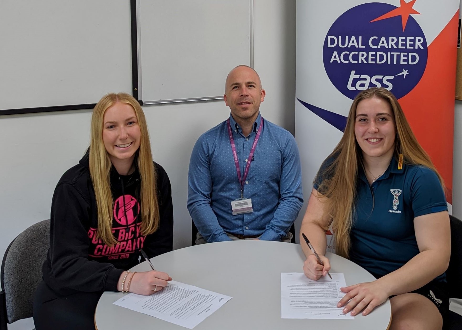 Two female stundents sign documents at table as man looks on