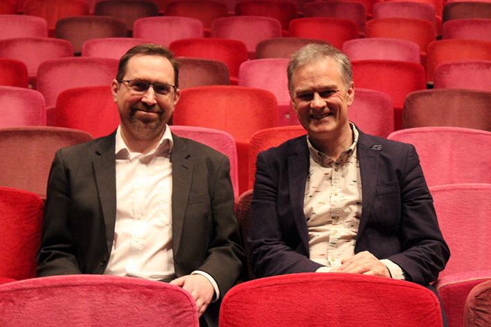 Two men sitting side by side on red velvet theatre seats