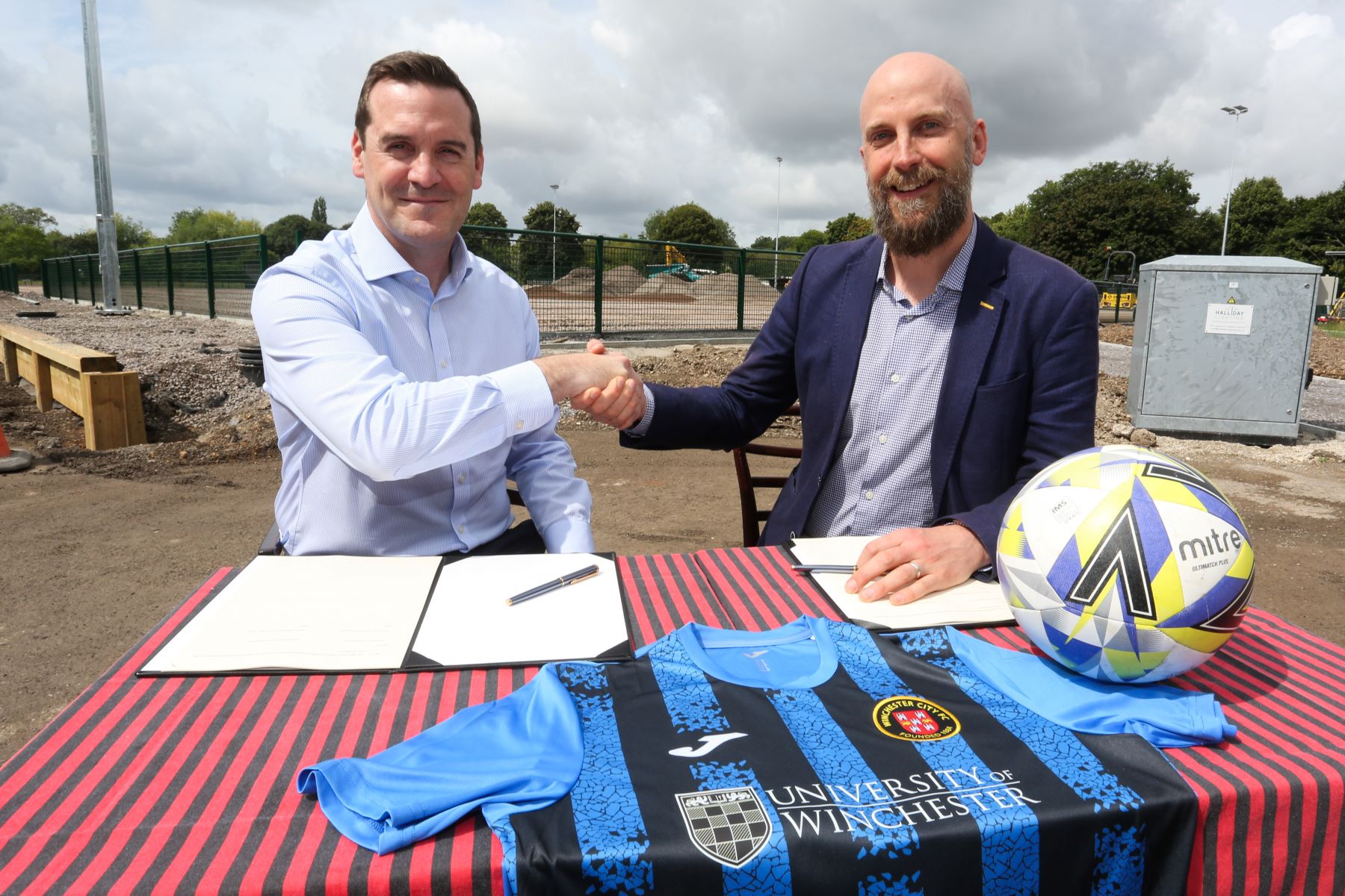 Two men shake hands while sat at a desk with football shirt and football