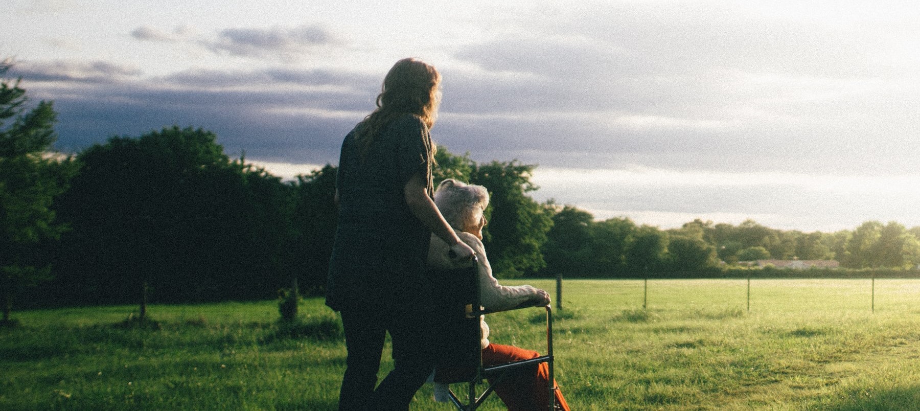 A young woman pushing an older woman in a wheelchair on the grass towards the sunlight