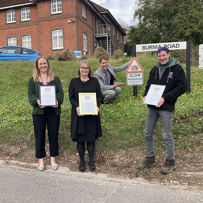 Four people holding gold certificates by a hedgehog road sign