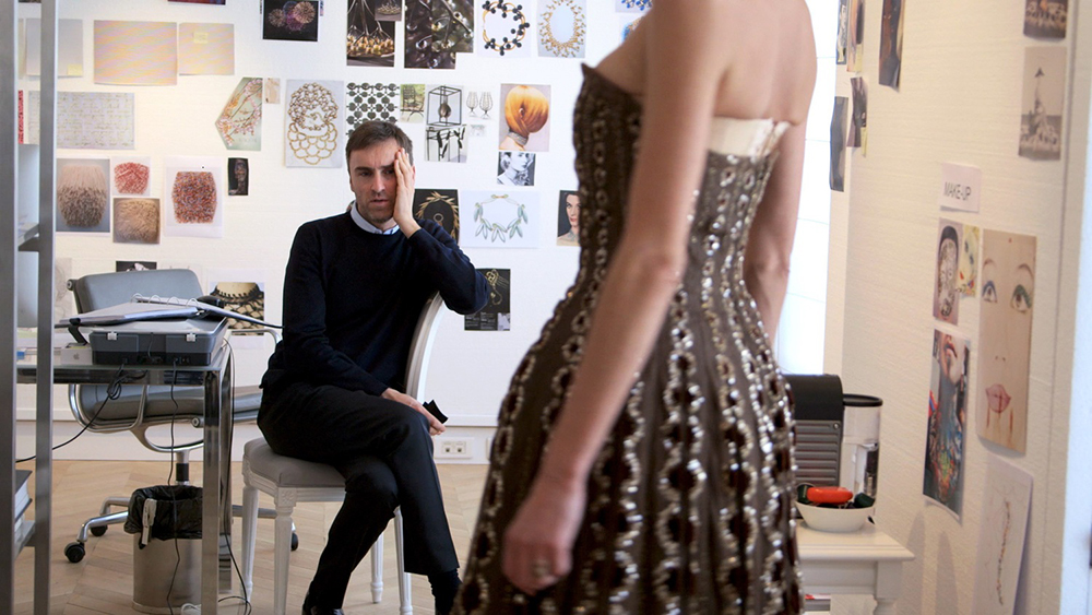 Man seated on chair observes woman in designer dress; design pictures on the walls