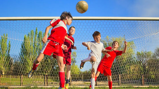 Four young boys playing football, one mid-air after headbutting the ball in front of the goal