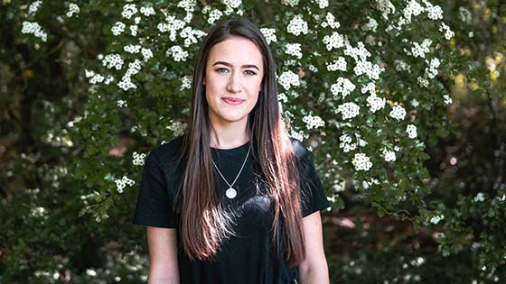 Hannah Lee studying for her Degree Apprenticeship, long brown hair smiling in front of white flowers