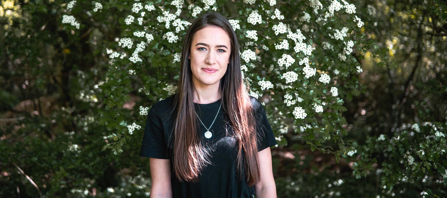 Young woman with long dark hair standing in front of the flowering hedge