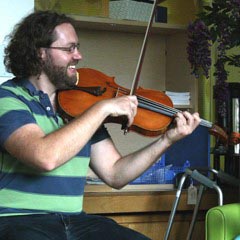 Man plays violin with elderly woman smiling playing percussion in chair