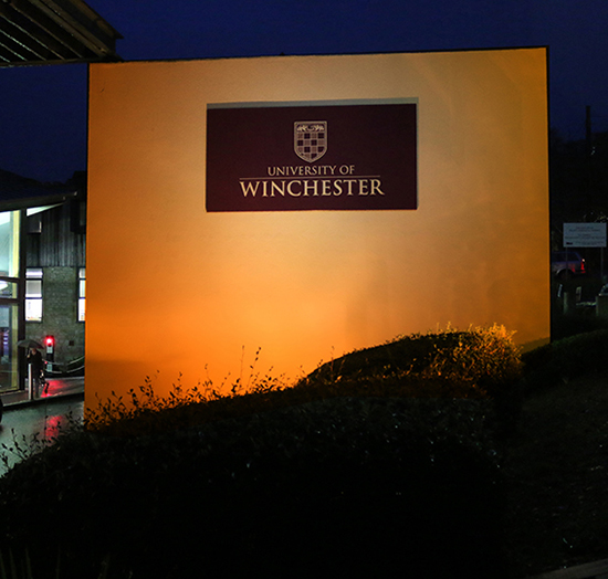 University of Winchester sign outside main reception lit in an orange glow at night