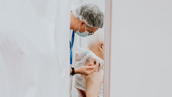 Male doctor in PPE administers vaccine into arm of man sitting down