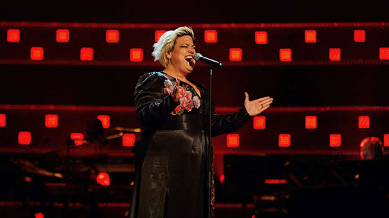 Hannah standing in front of a microphone with outstretched hands singing with dramatic black and red background