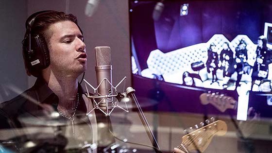 Student recording in sound booth sings into microphone while playing guitar