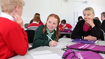 Two schoolchildren working at a desk watched by their female teacher