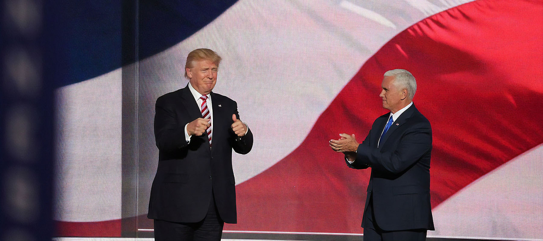 President Trump stands onstage clapping next to another ma against a backdrop of the stars and stripes flag