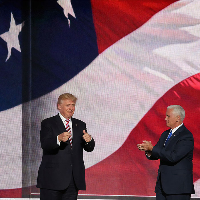 President Trump stands onstage clapping next to another ma against a backdrop of the stars and stripes flag