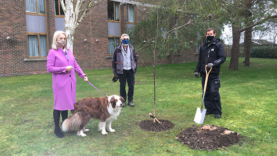 Joy carter in bright pink coat and with a dog on a lead standing with two male groundspeople next to a newly planted tree