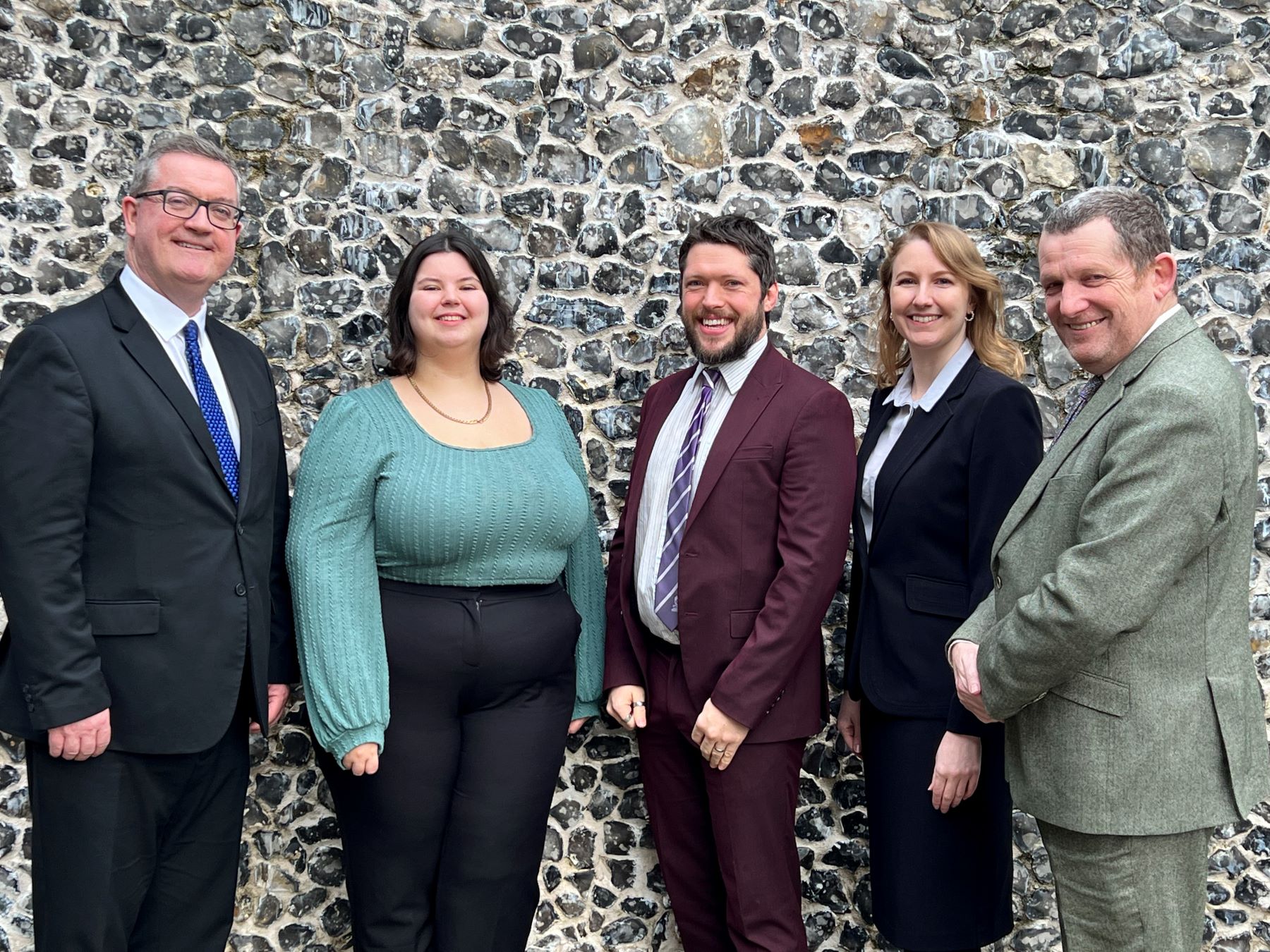 Five people in business attire, three men two women, in front of flinty wall