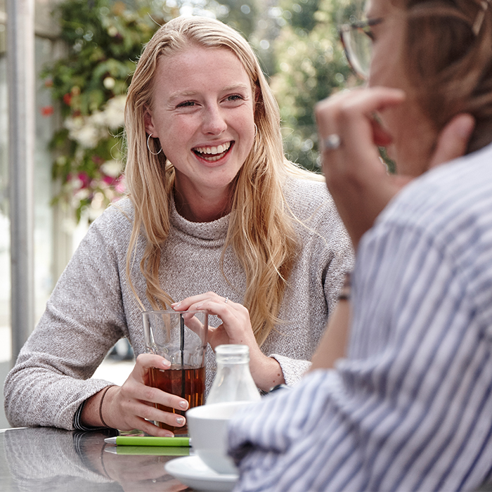 Female student sat at a coffee table smiling and having a conversation with a student to her right