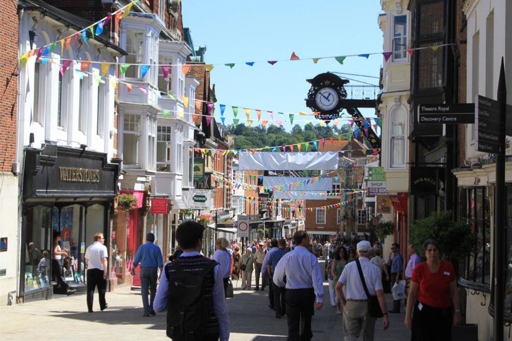 High street with bunting hanging across the rooftops, busy with people walking in the sun