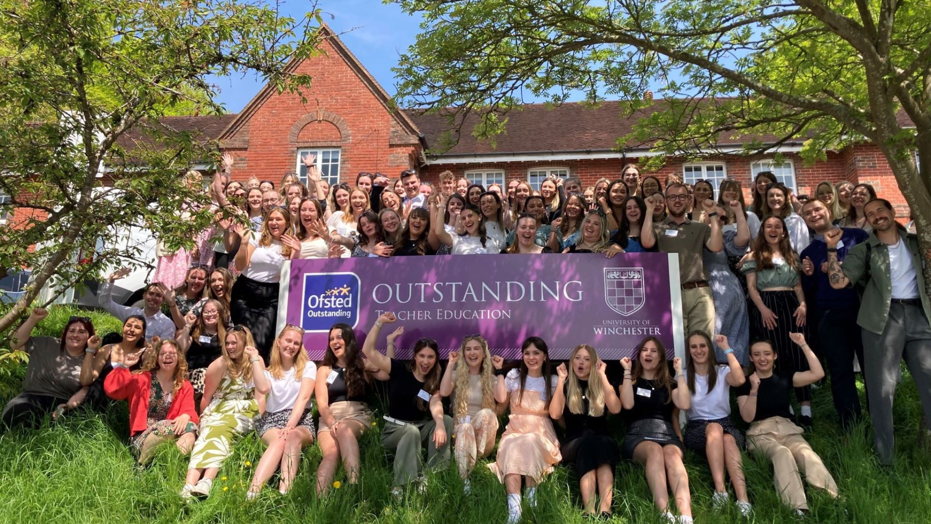 Teacher Training Students sitting under tree next to Ofsted Outstanding sign