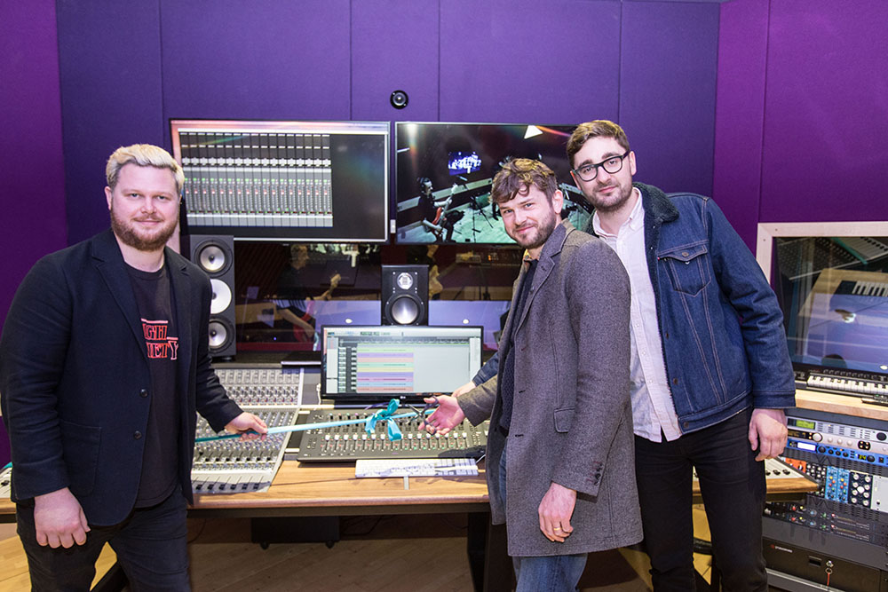 Three men standing by a mixing desk in a recording studio