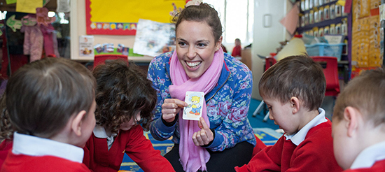 Young woman holding a card with cartoon bear and smiling at group of schoolchildren