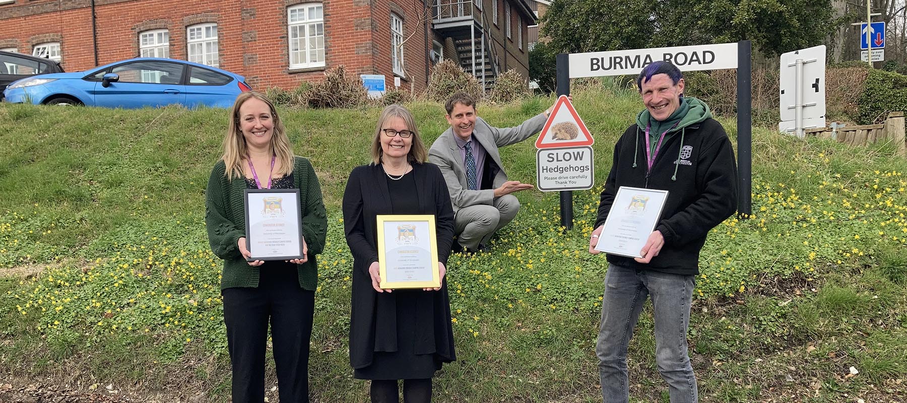 Four people holding gold certificates by a hedgehog road sign