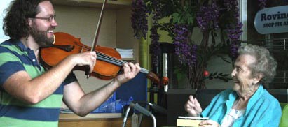 Man plays violin with elderly woman smiling playing percussion in chair