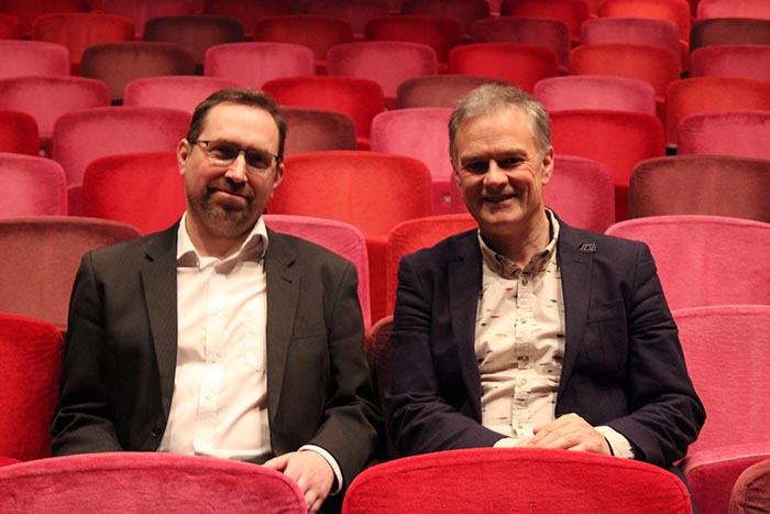 Two suited men smiling in red theatre seats